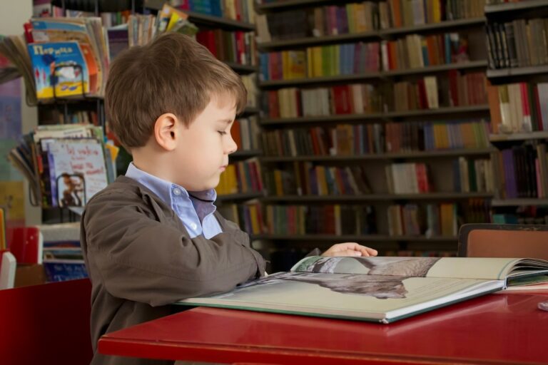 Young boy in a brown jacket reads a large open book at a red table in a library
