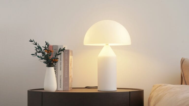 White mushroom lamp glowing on a round dark bedside table beside a vase and stacked books