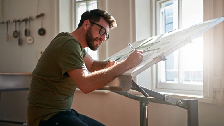 Man with glasses and beard sketches at an angled drafting table by a sunlit window