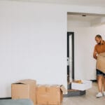 Man wearing an orange striped polo and shorts carries a large cardboard box in a doorway of a nearly empty living area with more boxes on the floor nearby