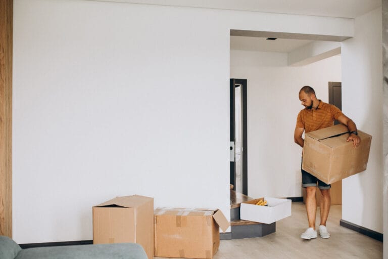 Man wearing an orange striped polo and shorts carries a large cardboard box in a doorway of a nearly empty living area with more boxes on the floor nearby