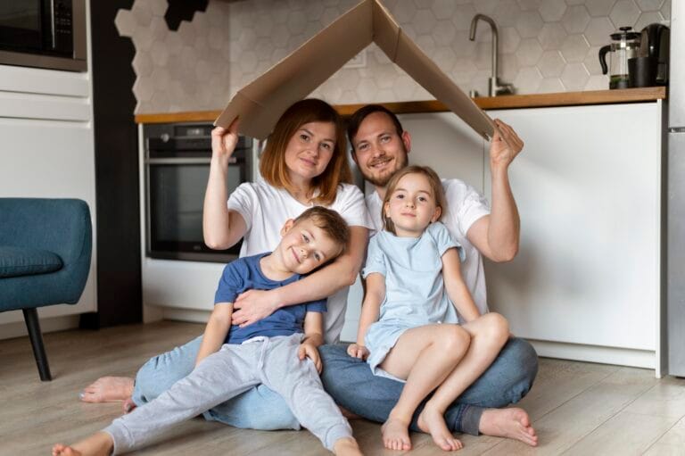 Family of four posing together on the floor under a cardboard roof in a modern kitchen smiling at the camera to create a cozy indoors moment