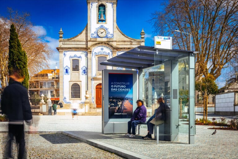 Historic church facade with blue azulejo tiles beside a modern glass bus shelter in a sunny urban square with pedestrians passing by