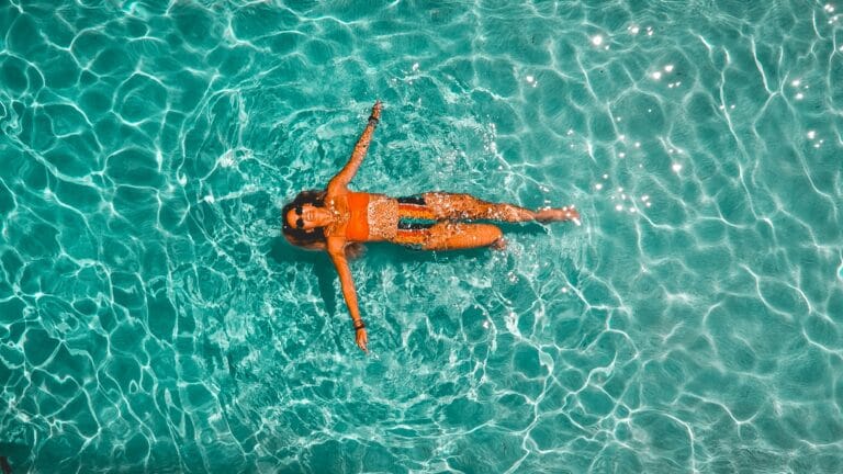 A person floating on their back in bright turquoise water wearing an orange swimsuit and sunglasses