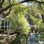 Round stone table with four matching stools under a large leafy tree in a garden beside a modern glass walled house and narrow pool