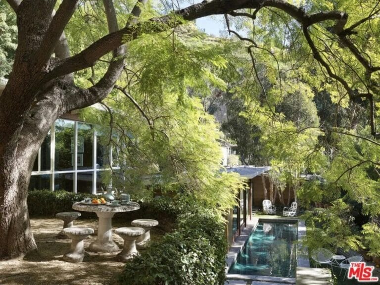Round stone table with four matching stools under a large leafy tree in a garden beside a modern glass walled house and narrow pool