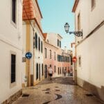 Narrow sunlit street between pastel buildings with orange tiled roofs a wrought iron lamp and pedestrians