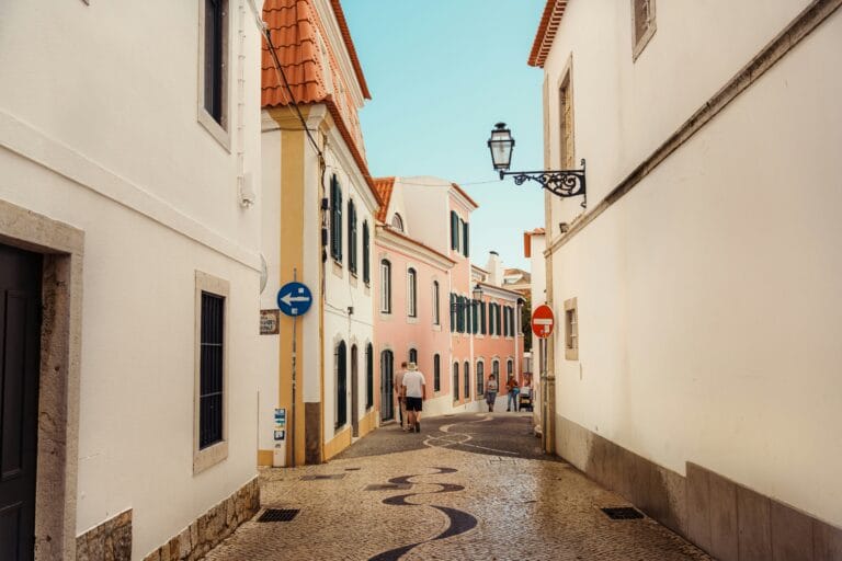 Narrow sunlit street between pastel buildings with orange tiled roofs a wrought iron lamp and pedestrians