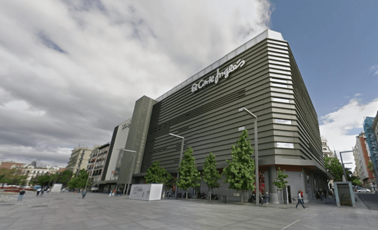Exterior of El Corte Inglés department store on a wide city plaza with trees and pedestrians nearby in a gray modern building setting