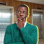 Man in a teal sweater with glasses stands in a hallway in front of two elevator doors posing as if thinking with his hand on his chin