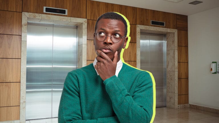 Man in a teal sweater with glasses stands in a hallway in front of two elevator doors posing as if thinking with his hand on his chin