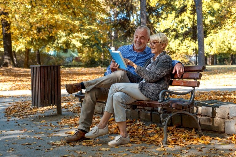 Elderly couple reading a blue book together on a park bench amid autumn leaves and sunlight