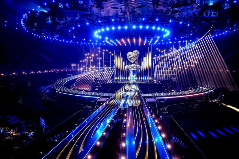 Elaborate Eurovision stage with blue lighting a circular overhead light ring and glowing ramps leading to a central performance area marked Eurovision Song Contest