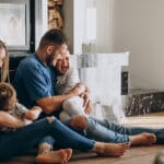 Man smiles while hugging a laughing child two more kids sit nearby on a wooden floor by a lit fireplace in a cozy living room