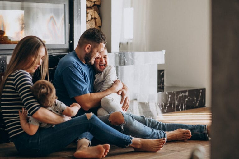 Man smiles while hugging a laughing child two more kids sit nearby on a wooden floor by a lit fireplace in a cozy living room