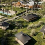 Aerial view of a campground with black canvas tents wooden cabins a rectangular pool a basketball court and grassy paths among trees