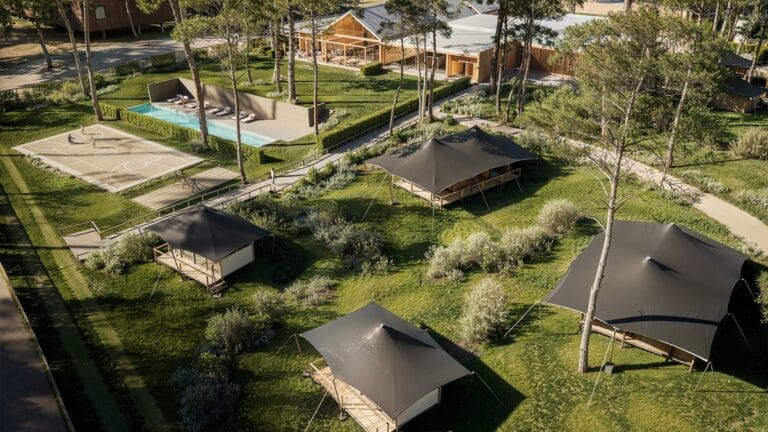 Aerial view of a campground with black canvas tents wooden cabins a rectangular pool a basketball court and grassy paths among trees