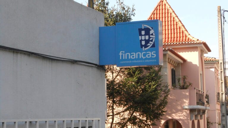 Blue sign reading financas with a circular logo mounted on a blue box along a street with pink houses in the background
