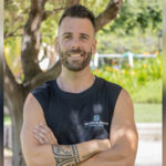 Smiling man with short dark hair and beard arms crossed wearing a black sleeveless shirt in a sunny park tattooed forearm visible