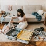 Woman kneels on a rug folding clothes to pack into a large yellow and gray suitcase in a bright living room with a sofa and scattered garments nearby