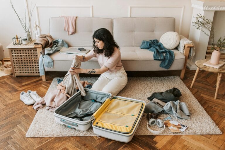 Woman kneels on a rug folding clothes to pack into a large yellow and gray suitcase in a bright living room with a sofa and scattered garments nearby