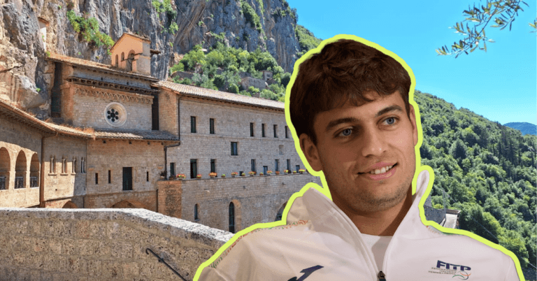 Smiling young man in a white jacket posed in front of a stone monastery set against greenery and hills on a sunny day