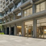 Modern concrete office building with large glass storefronts and a paved plaza with people walking and talking outside