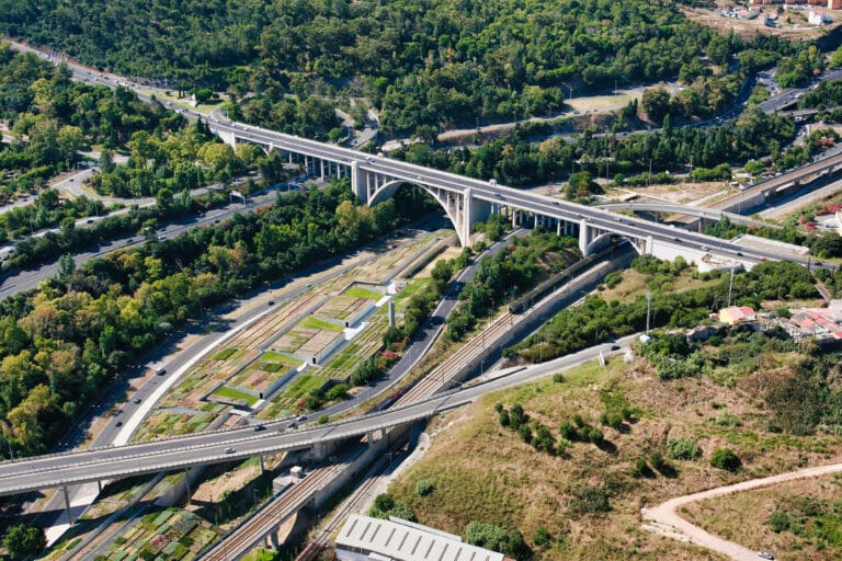 Aerial view of a sprawling highway interchange with tall concrete bridges curved ramps and surrounding greenery and train tracks nearby