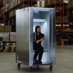 Woman sits inside a tall silver metal cabinet on wheels in a warehouse aisle