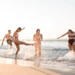 Group of friends in swimsuits playing in shallow ocean water at the beach as waves splash around them during sunset