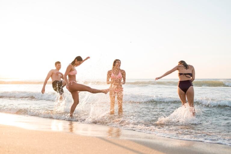 Group of friends in swimsuits playing in shallow ocean water at the beach as waves splash around them during sunset
