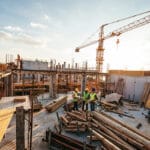 Construction site scene with a large crane overhead concrete forms and workers in hard hats and reflective vests nearby