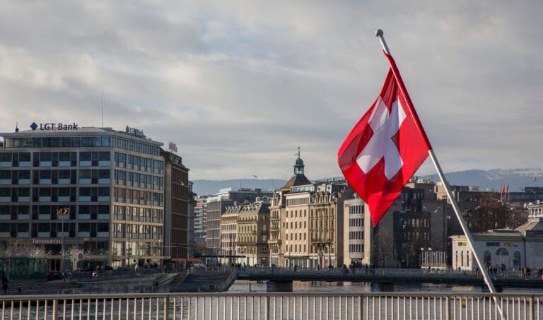 Swiss flag waving on a tall pole over a riverfront cityscape with modern buildings and a cloudy sky