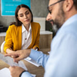 Woman in a yellow blazer sits at a desk as a colleague hands her documents in an office