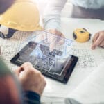 Engineers review blueprints on a drafting table with a yellow hard hat and a tablet showing a 3D building model