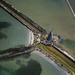 Aerial view of a stone windmill on a small islet connected by breakwaters to the shore with turquoise water and sandy shores around
