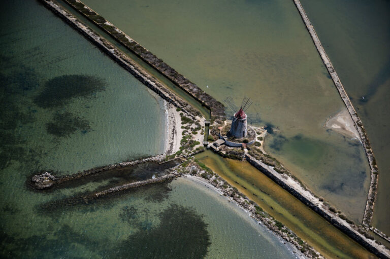 Aerial view of a stone windmill on a small islet connected by breakwaters to the shore with turquoise water and sandy shores around