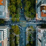 Aerial view of a tree lined city street crossing between tall buildings with cars and crosswalks visible below the sidewalks