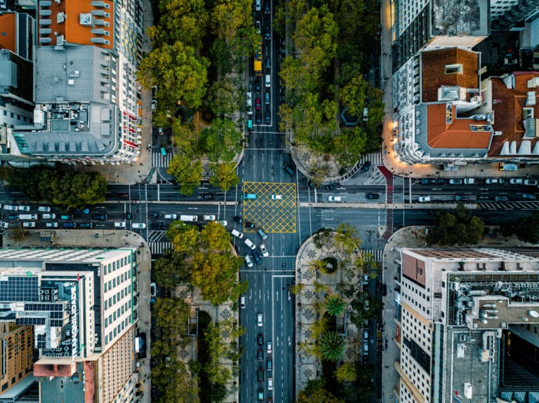 Aerial view of a tree lined city street crossing between tall buildings with cars and crosswalks visible below the sidewalks