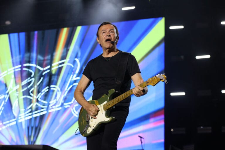 Male guitarist performing on stage with a green electric guitar in front of a colorful abstract LED backdrop