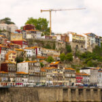 Colorful hillside row of houses above a stone riverwall with a tall construction crane in the background under a cloudy sky