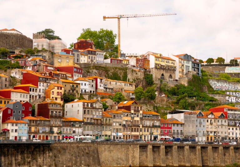 Colorful hillside row of houses above a stone riverwall with a tall construction crane in the background under a cloudy sky