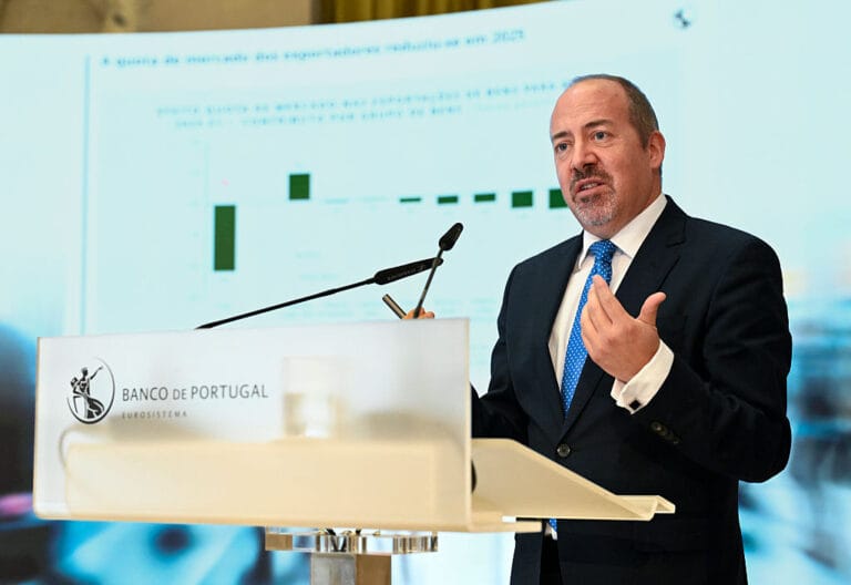 Man in a dark suit and blue tie speaks at a Banco de Portugal podium with a microphone and a presentation slide behind him