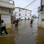 People wading through a flooded street carrying bags and containers amid submerged buildings and parked cars off to the side in a town center
