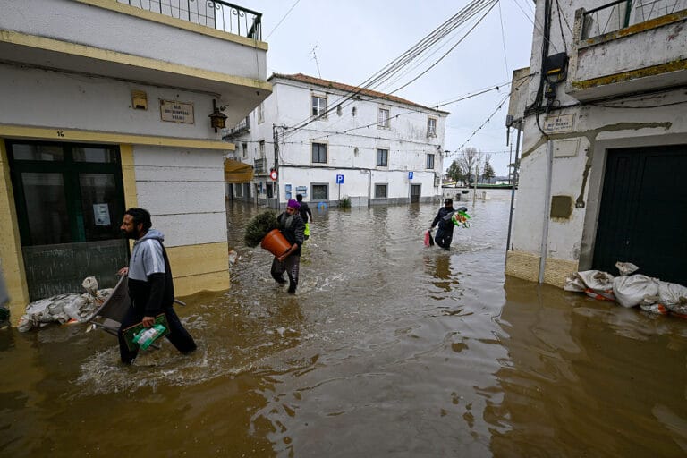 People wading through a flooded street carrying bags and containers amid submerged buildings and parked cars off to the side in a town center