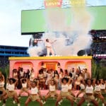 Dancers in white outfits perform on a pink stage with smoke in a packed stadium and a huge scoreboard above them