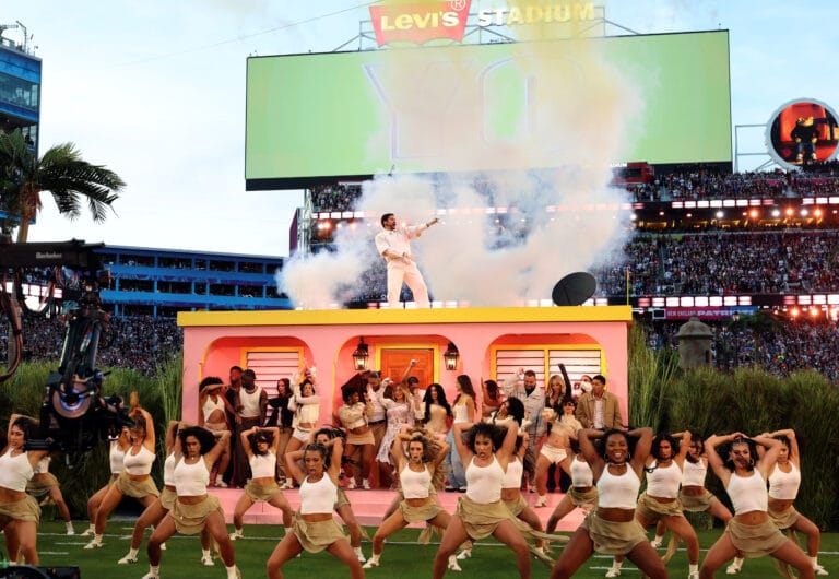 Dancers in white outfits perform on a pink stage with smoke in a packed stadium and a huge scoreboard above them
