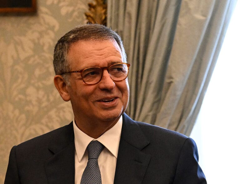 Older man in a dark suit and tie wearing brown glasses smiling in a formal room with patterned wallpaper and drapes