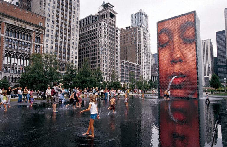 Children splash in a shallow reflecting pool at Crown Fountain in Millennium Park with a giant red brick video tower spouting water from a large face image and city buildings in the background