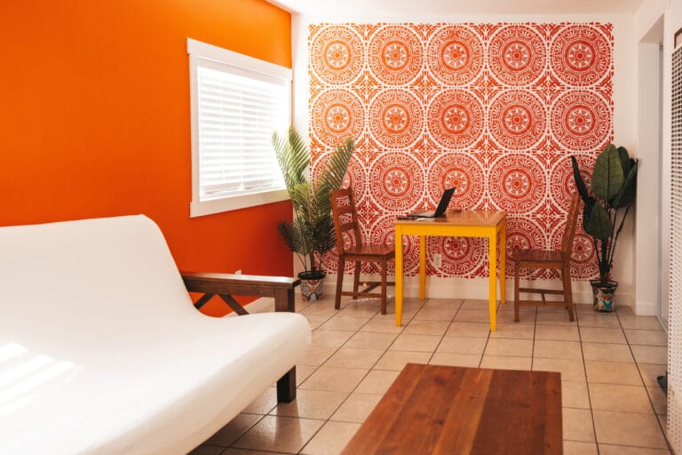 Bright orange wall with a white window blind and a red circular patterned wallpapered wall with a yellow table and chairs nearby Plants flank the scene